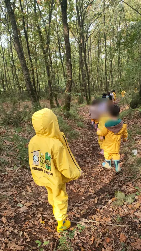 Sırtında Orman Okulu logolu sarı yağmurluk olan çocuk, arkadaşlarının ardından orman patikasında yürüyor